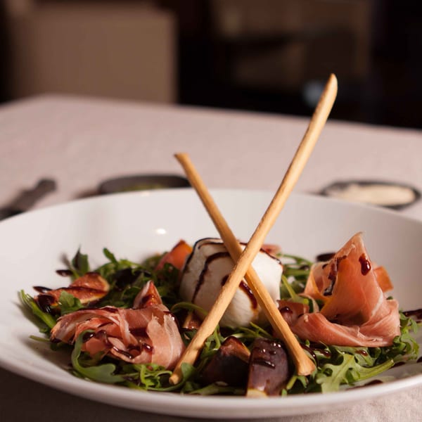 Arugula salad with prosciutto, figs, mozzarella, balsamic glaze, and breadsticks in a white bowl.