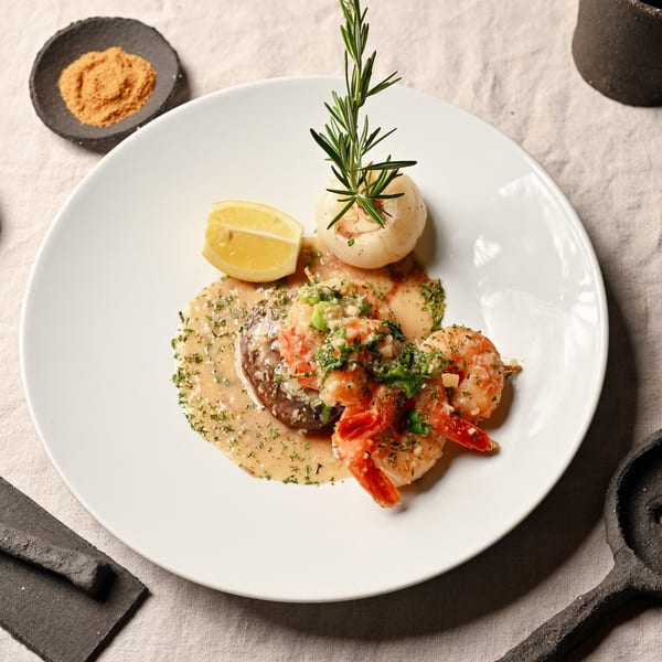 Gourmet dish with shrimp, steak, lemon, and a mushroom garnished with rosemary on a white plate.