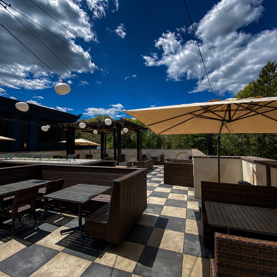 Outdoor patio with checkered floor, dark wooden booths, beige umbrellas, and string lights under a blue sky.