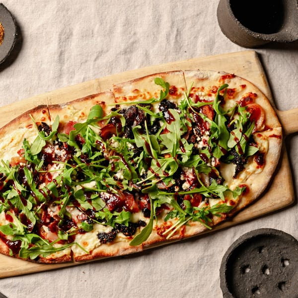 Flatbread pizza with arugula, cheese, bacon, figs, and balsamic glaze on a wooden board.