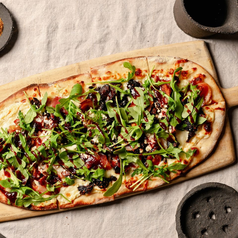 Flatbread pizza with arugula, cheese, bacon, figs, and balsamic glaze on a wooden board.