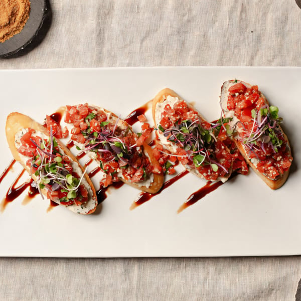 Five bruschetta pieces with diced tomatoes, microgreens, and balsamic glaze on a white platter.